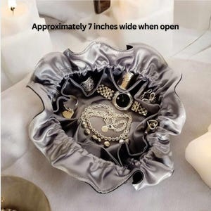 Op de afbeelding: Een grijze satijnen juwelendoos met een trekkoord. De doos is open en gevuld met sieraden, waaronder een horloge, kettingen en ringen. De doos is ongeveer 18 cm breed wanneer deze open is.