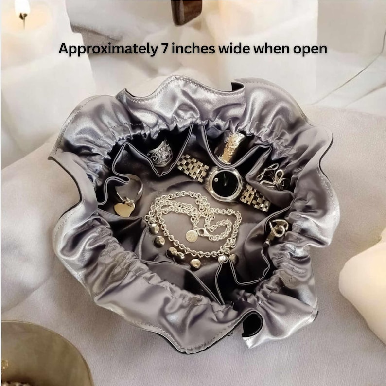 Op de afbeelding: Een grijze satijnen juwelendoos met een trekkoord. De doos is open en gevuld met sieraden, waaronder een horloge, kettingen en ringen. De doos is ongeveer 18 cm breed wanneer deze open is.