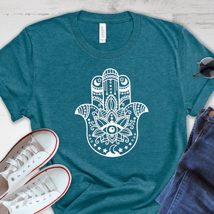 May include: A teal blue t-shirt with a white hamsa hand design. The hamsa hand has a stylized eye in the center and is surrounded by intricate details. The t-shirt is laid flat on a white wooden surface with a pair of white sneakers and blue jeans.
