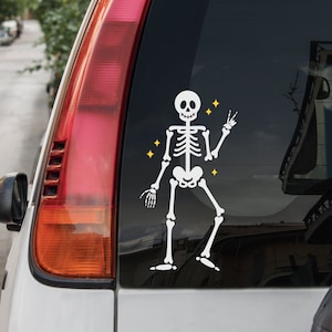 May include: White skeleton sticker with a peace sign on a black car window. The skeleton is wearing a white shirt and has yellow stars around it.