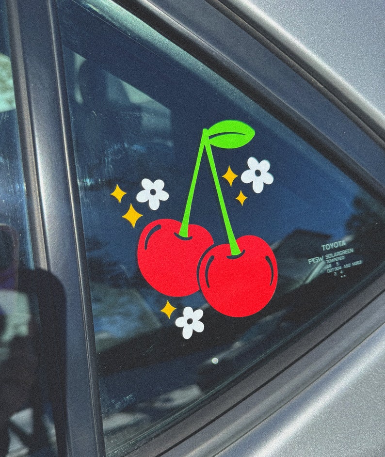 May include: A red cherry sticker with a green stem and leaves, two white flowers, and gold glitter stars on a car window.