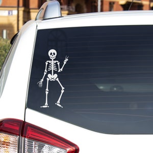 May include: A white car with a skeleton sticker on the back window. The skeleton is waving with one hand and has a friendly expression.
