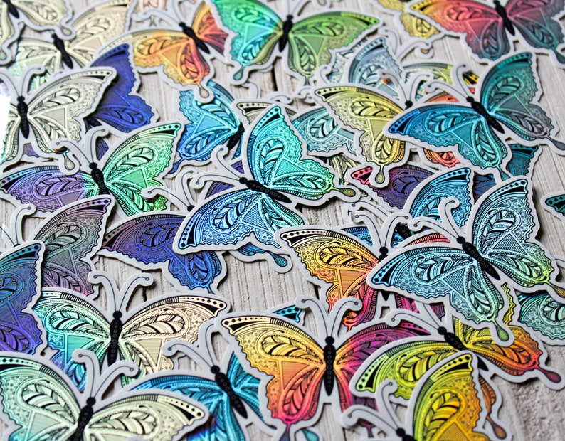 May include: A collection of colorful butterfly stickers with intricate patterns. The stickers feature a variety of colors, including blue, green, yellow, and purple, with a metallic sheen. The butterflies have detailed wing designs and are arranged on a light-colored surface.