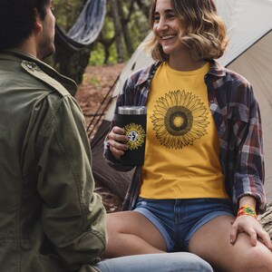 May include: A black tumbler with a sunflower design and initials, held by a person wearing a yellow sunflower t-shirt and denim shorts. The scene is set outdoors with a tent and trees in the background.
