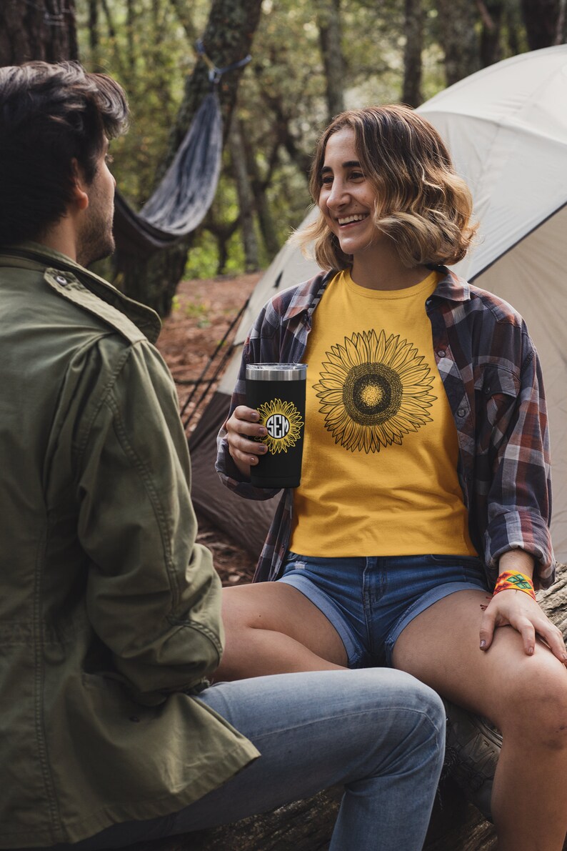 May include: A black tumbler with a sunflower design and initials, held by a person wearing a yellow sunflower t-shirt and denim shorts. The scene is set outdoors with a tent and trees in the background.