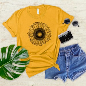 May include: Mustard yellow t-shirt with a black sunflower graphic. The shirt is paired with denim shorts, sunglasses, and a vintage camera. A large green leaf adds a natural element to the composition.