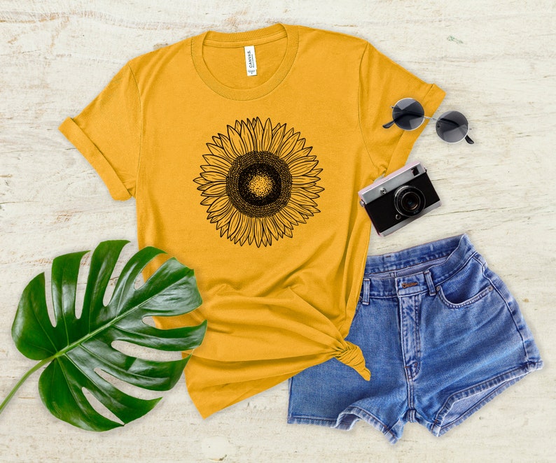 May include: Mustard yellow t-shirt with a black sunflower graphic. The shirt is paired with denim shorts, sunglasses, and a vintage camera. A large green leaf adds a natural element to the composition.