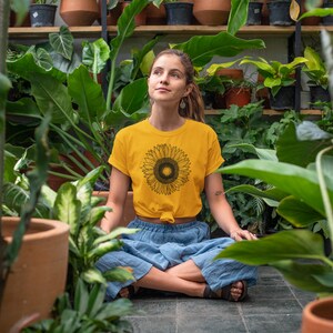May include: A yellow t-shirt with a black sunflower graphic. The person is wearing blue pants and is surrounded by lush green plants in a greenhouse setting. The shirt is knotted at the waist.