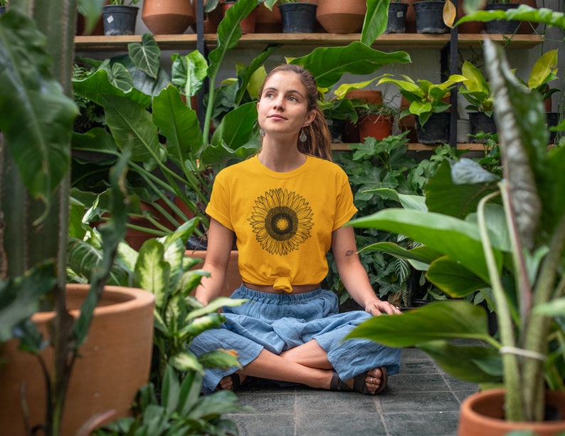 May include: A yellow t-shirt with a black sunflower graphic. The person is wearing blue pants and is surrounded by lush green plants in a greenhouse setting. The shirt is knotted at the waist.
