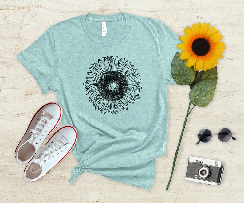 May include: A light blue t-shirt featuring a black and white sunflower graphic. The shirt is styled with white sneakers, a yellow sunflower, sunglasses, and a vintage camera. The shirt is knotted at the waist.