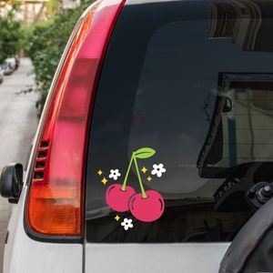 May include: A car window decal with two red cherries and white flowers. The cherries have green stems and leaves.