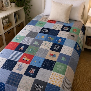 May include: A patchwork quilt featuring various blue, red, and grey squares with printed graphics of rockets, trucks, bears, and text including "Little Brother," "Awesome," "Born to be Wild," "Cool Kid," and "Little Dude" on a single bed.
