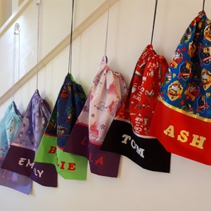 Pode incluir: Seis sacos de cord&atilde;o de cores diferentes pendurados em ganchos. Cada saco tem um design diferente e um nome bordado na parte inferior: Lily, Emily, Billie, Isla, Tom e Ash.