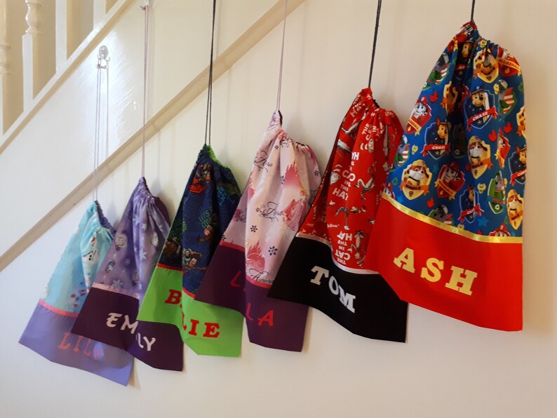 Pode incluir: Seis sacos de cord&atilde;o de cores diferentes pendurados em ganchos. Cada saco tem um design diferente e um nome bordado na parte inferior: Lily, Emily, Billie, Isla, Tom e Ash.