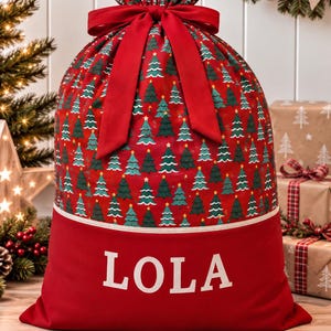 May include: A red fabric gift sack featuring a repeating pattern of green Christmas trees with yellow stars. The top is cinched with a red ribbon bow. The bottom section is solid red with the name LOLA printed in large white capital letters.