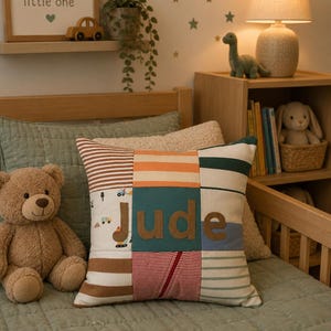 May include: A patchwork cushion with the name "Jude" in brown letters. The cushion features various fabric patterns, including stripes and vehicle prints. A teddy bear sits beside the cushion on a bed with a green quilt.