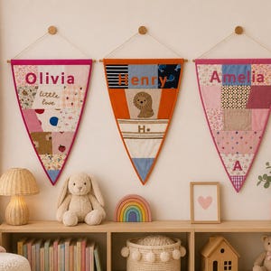 May include: Three triangular fabric wall banners hanging in a row. The left banner is pink with the name Olivia and the text little love. The centre banner is orange with the name Henry and a lion graphic. The right banner is pink with the name Amelia.