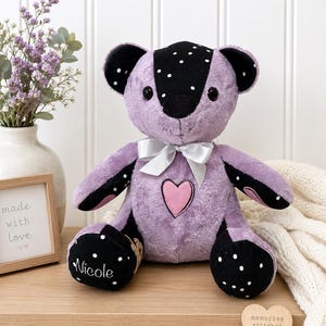 May include: A plush teddy bear featuring lavender and black fabric with white polka dots. The bear has a pink heart patch on its chest, a white satin ribbon around its neck, and the name Nicole embroidered in white on one of its black paws.