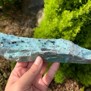 May include: A piece of raw, natural stone with a light blue and green mineral surface. The stone has dark flecks and a rough, textured appearance. The edges are a darker gray color. The stone is held in a hand.