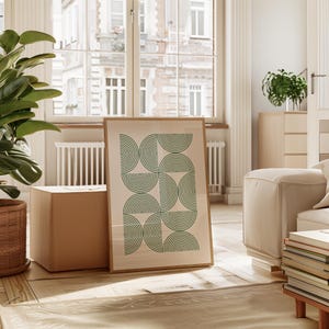 May include: A framed print with a geometric design of green concentric circles on a white background. The print is leaning against a cardboard box in a room with a large window and a white couch.