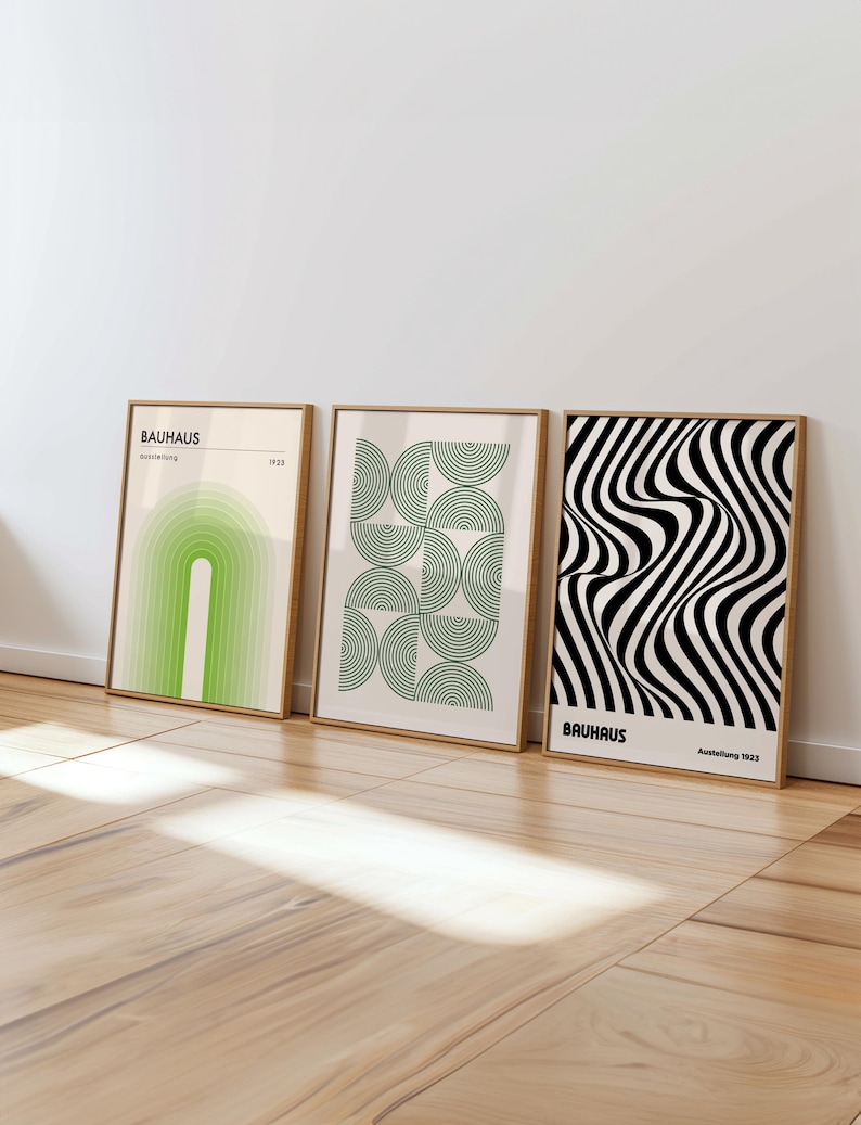 Three framed Bauhaus-style posters leaning against a white wall on a light wooden floor. The left poster features a green arch-like gradient pattern with the text “BAUHAUS ausstellung 1923.” The center poster shows a grid of green concentric semicircles forming a geometric design. The right poster displays a bold black and white optical illusion pattern of wavy lines with the text “BAUHAUS Ausstellung 1923.
