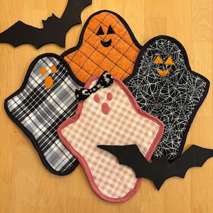May include: A collection of Halloween-themed pot holders and decorative bats. The pot holders are ghost-shaped with various patterns, including plaid, spiderwebs, and gingham. The bats are black. The pot holders have smiling faces.