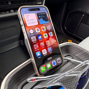 Puede incluir: Un iPhone blanco con una pantalla negra que muestra la hora 10:17, la fecha sábado 22 y varios iconos de aplicaciones. El teléfono está en un soporte de teléfono para coche, y un par de gafas de sol transparentes con lentes azules descansan sobre el soporte del teléfono.