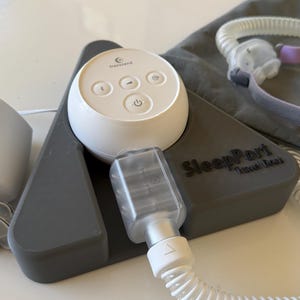 Puede incluir: Un dispositivo blanco y gris para la apnea del sueño con el texto "SleepPort Travel Dock". El dispositivo incluye un panel de control blanco con botones y un componente de plástico transparente conectado por una manguera blanca. También se ve una funda de transporte gris.
