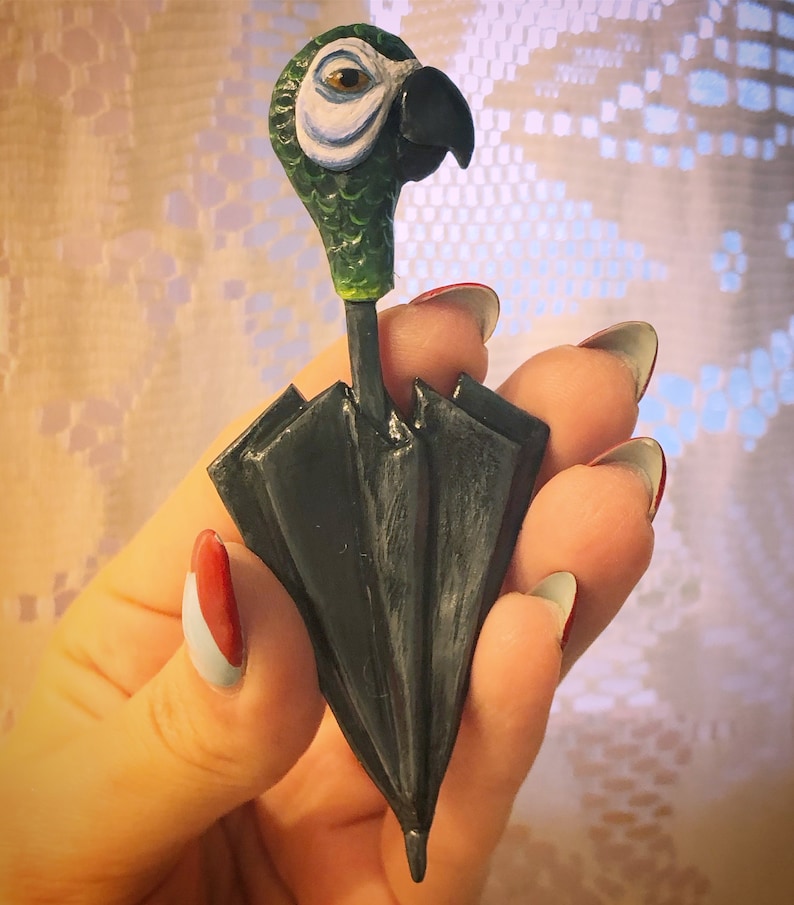 May include: A green and blue parrot head figurine attached to a black umbrella-shaped handle. The figurine is made of painted ceramic and is a whimsical and decorative piece.