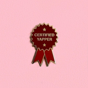 May include: A maroon and gold enamel pin in the shape of a ribbon with the words "CERTIFIED YAPPER" in gold lettering. The pin has two small stars and a red ribbon design, set against a pink background.