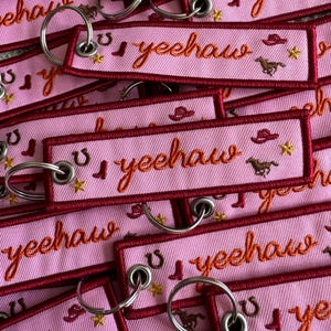 May include: Pink embroidered keyrings with the word "yeehaw" in orange script. Each keyring features a red border and small embroidered images of cowboy boots, horseshoes, cowboy hats, horses, and stars. Silver key rings are attached.