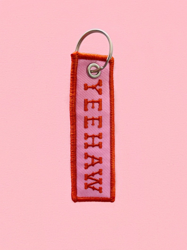 May include: A pink fabric keychain with an orange embroidered "YEEHAW" on it. The keychain has a silver metal ring.