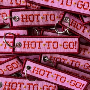May include: A close-up of several pink fabric keychains with the text "HOT TO GO!" embroidered in orange and yellow thread. The keychains are stacked on top of each other, and the text is visible on each keychain.