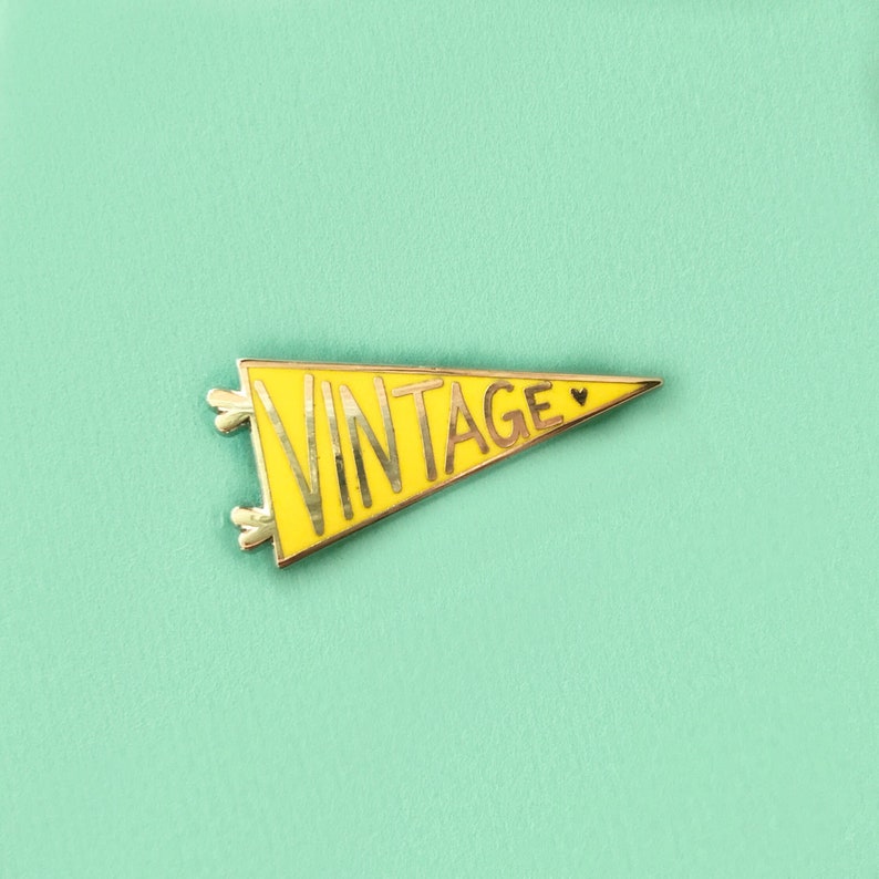 May include: A yellow pennant-shaped pin with the word "VINTAGE" in gold lettering. The pin has a gold border and small decorative elements at the base. The pin is set against a light teal background.