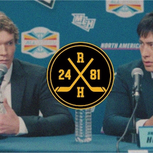 May include: A black and gold hockey logo with crossed hockey sticks, the numbers 24 and 81, and the letters R and H. The logo is centered in front of two people wearing suits and speaking into microphones.