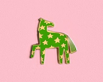 May include: A green horse-shaped enamel pin with a gold outline and gold star pattern. The pin has a gold mane and tail, set against a pink background. This decorative pin is a fun accessory.