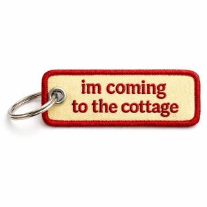 May include: A rectangular keychain with the phrase "im coming to the cottage" embroidered in red on a cream-colored background. The keychain has a red border and a silver key ring.