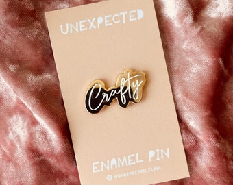May include: Gold enamel pin with the word "Crafty" in white lettering on a beige card with the text "UNEXPECTED" and "ENAMEL PIN" in white lettering. The card also includes the text "@UNEXPECTED_FLAIR".