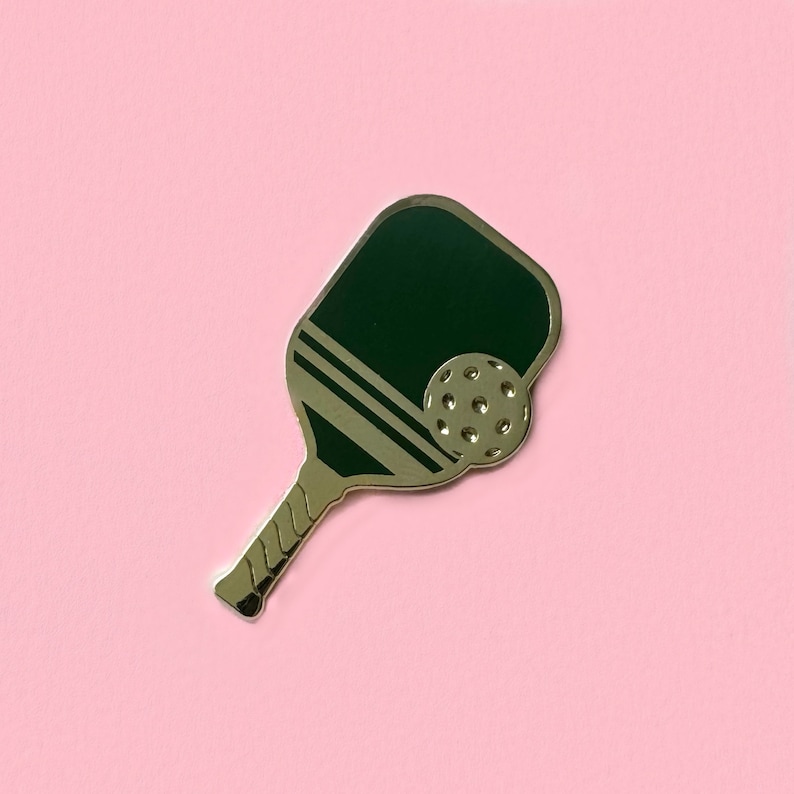 Op de afbeelding: Een gouden pickleball-bat met een groen oppervlak en een witte streep. De bat heeft een ronde, witte bal met putjes.