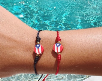 May include: Two bracelets with the Puerto Rican flag design. One bracelet is black, the other is red. Each bracelet has a round charm with the flag's colors: red, white, and blue. The bracelets are on a wrist near a pool.