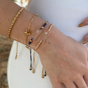 May include: A collection of gold bracelets adorns a wrist. The bracelets include a thick rope chain, a delicate chain with a star charm, and several beaded and corded designs. The bracelets are layered and create a stylish, elegant look.