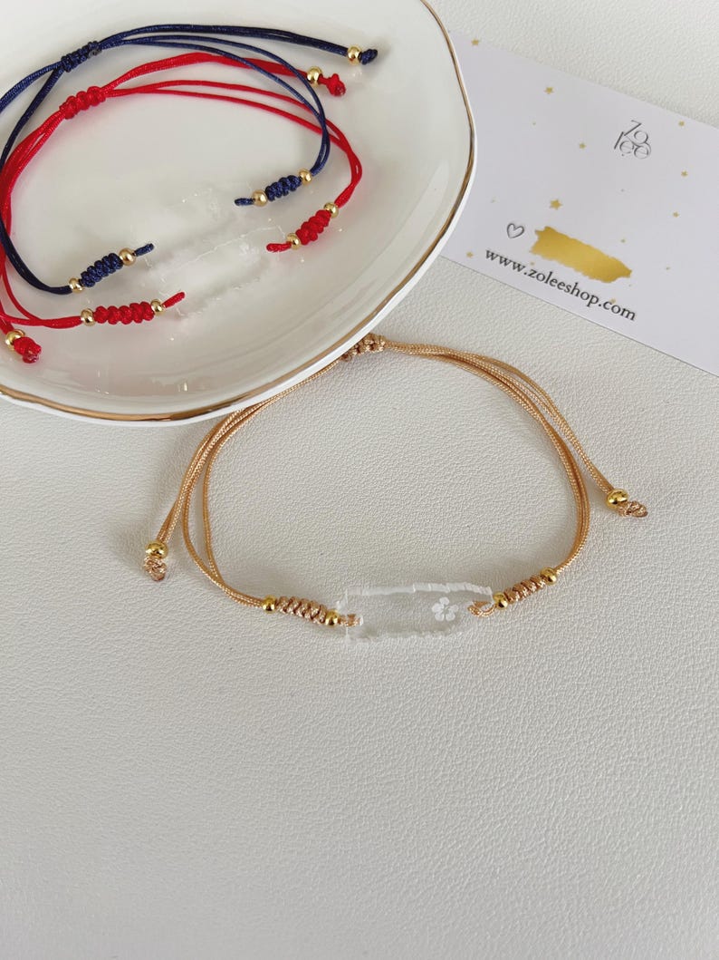 May include: A collection of handmade bracelets. The bracelets feature red, navy blue, and gold cord with gold-colored beads. One bracelet has a clear rectangular charm with a floral design. The website www.zoleeshop.com is visible.