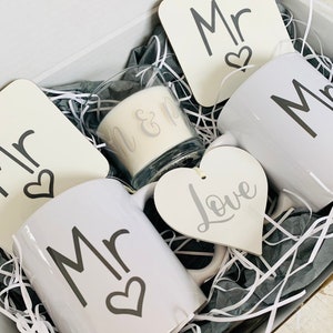 May include: A gift box filled with white mugs, coasters, and a candle. The mugs and coasters have the word "Mr." and a heart design. The candle has the words "Mr. & Mrs." on it. A white heart-shaped ornament with the word "Love" is also included.