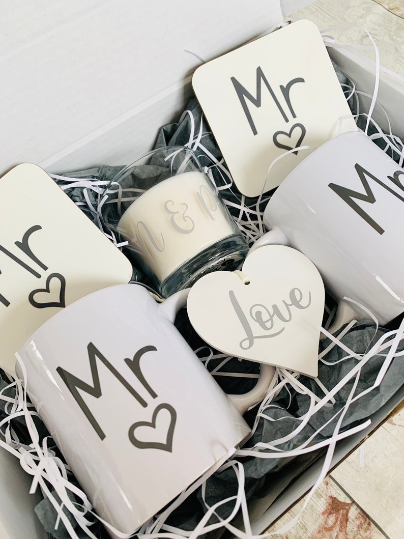 May include: A gift box filled with white mugs, coasters, and a candle. The mugs and coasters have the word "Mr." and a heart design. The candle has the words "Mr. & Mrs." on it. A white heart-shaped ornament with the word "Love" is also included.