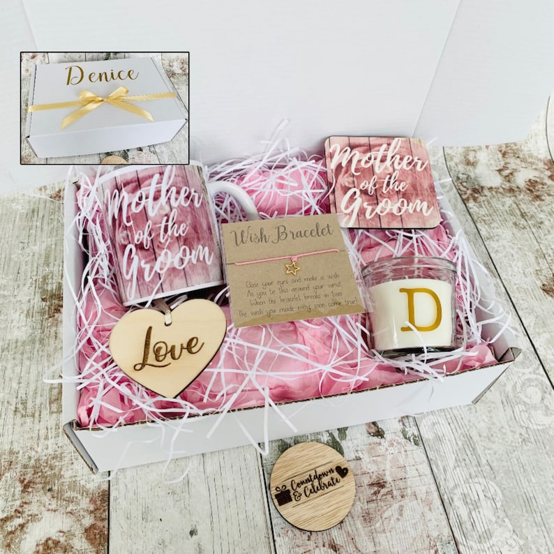 May include: A white gift box filled with a pink and white shredded paper. The box contains a mug that says "Mother of the Groom", a wooden heart that says "Love", a small candle with a gold letter "D", a wish bracelet, and two wooden coasters that say "Mother of the Groom" and "Countdown & Celebrate".