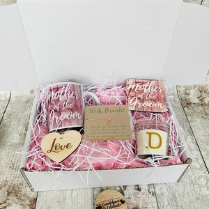 May include: A white gift box filled with pink tissue paper and a variety of gifts for a mother of the groom. The gifts include a mug, a coaster, a heart-shaped wooden sign, a candle, and a wish bracelet. The mug and coaster both feature the text "Mother of the Groom". The heart-shaped wooden sign says "Love". The candle is a clear glass jar with a white wax candle inside and a gold letter "D" on the front. The wish bracelet is a thin string with a gold star charm. The box also includes a small wooden circle with the text "Countdown & Celebrate".