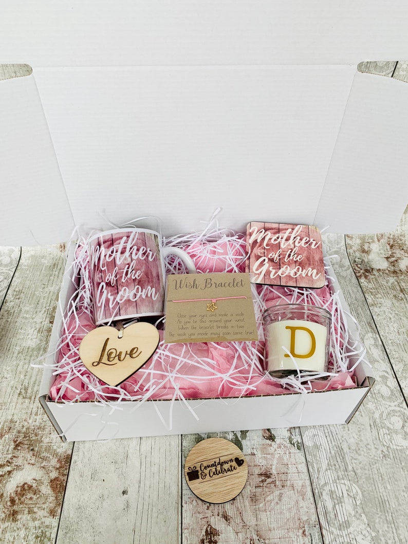 May include: A white gift box filled with pink tissue paper and a variety of gifts for a mother of the groom. The gifts include a mug, a coaster, a heart-shaped wooden sign, a candle, and a wish bracelet. The mug and coaster both feature the text "Mother of the Groom". The heart-shaped wooden sign says "Love". The candle is a clear glass jar with a white wax candle inside and a gold letter "D" on the front. The wish bracelet is a thin string with a gold star charm. The box also includes a small wooden circle with the text "Countdown & Celebrate".