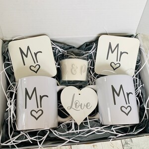 May include: A gift box filled with white shredded paper and a wedding gift set. The set includes two white mugs with the word "Mr" and a heart, two white coasters with the word "Mr" and a heart, a white heart-shaped ornament with the word "Love", and a white candle with the words "Mr & Mr" on it.