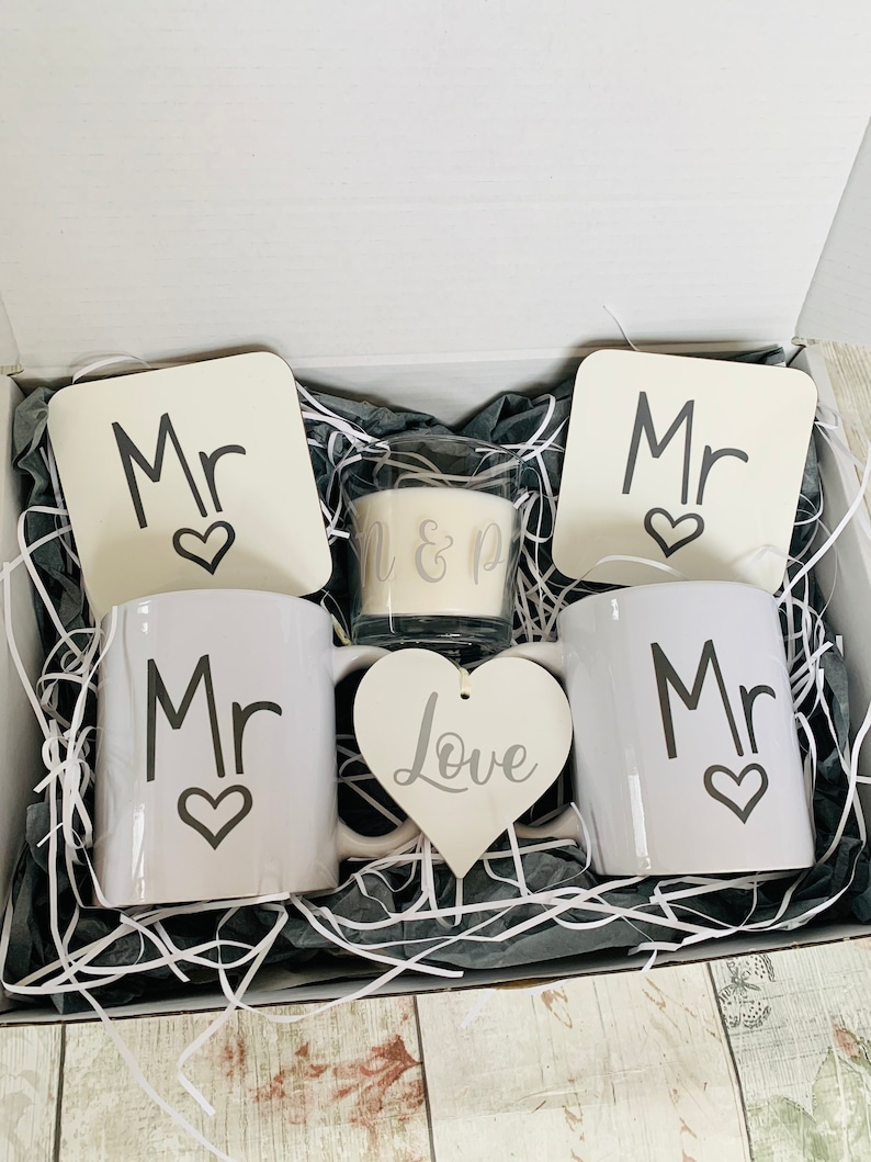 May include: A gift box filled with white shredded paper and a wedding gift set. The set includes two white mugs with the word "Mr" and a heart, two white coasters with the word "Mr" and a heart, a white heart-shaped ornament with the word "Love", and a white candle with the words "Mr & Mr" on it.