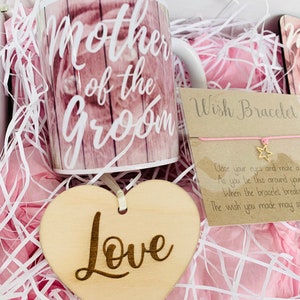 May include: A gift box filled with pink tissue paper and a white mug with the text "Mother of the Groom" printed on it. The box also contains a wooden heart with the word "Love" engraved on it and a wish bracelet with a star charm.